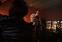 An elderly woman dances joyfully in a lively bar setting with friends and a bartender.