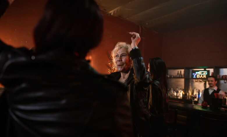 An elderly woman dances joyfully in a lively bar setting with friends and a bartender.