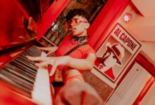 A man in a crop top with curly hair plays piano in an artful, vibrant setting.