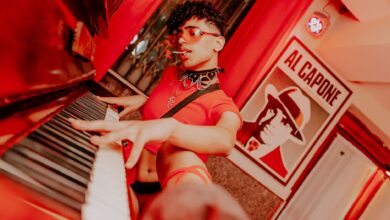 A man in a crop top with curly hair plays piano in an artful, vibrant setting.