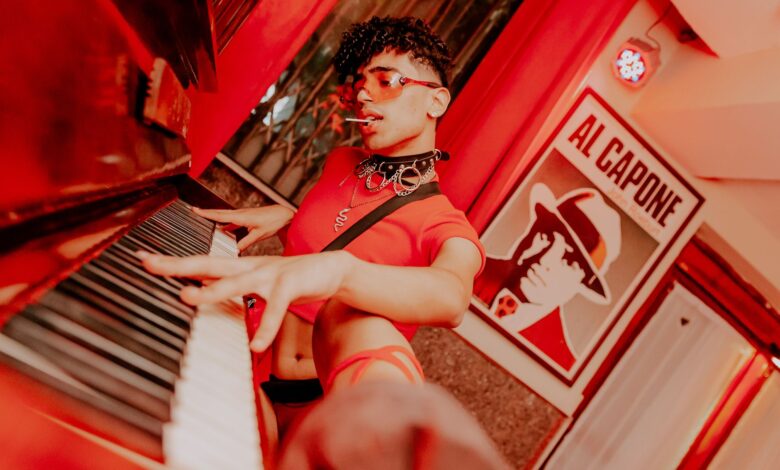 A man in a crop top with curly hair plays piano in an artful, vibrant setting.