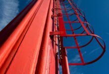 Dramatic low angle view of a red metal ladder on an industrial chimney against a clear blue sky.