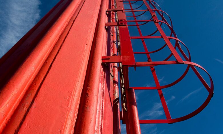 Dramatic low angle view of a red metal ladder on an industrial chimney against a clear blue sky.