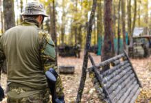 A man wearing camouflage gear participates in an outdoor paintball game in a forested area.