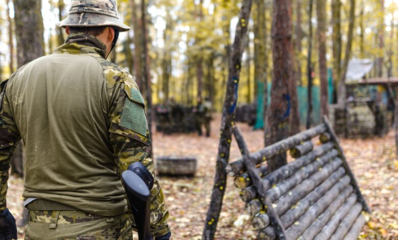 A man wearing camouflage gear participates in an outdoor paintball game in a forested area.