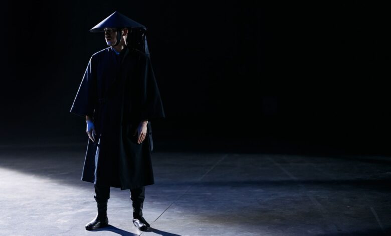 Dramatic studio portrait of a person in a dark costume with shadowed lighting.