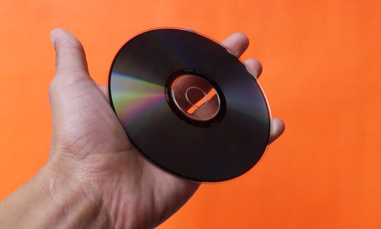 Close-up of a hand holding a classic compact disc with an orange background, evoking nostalgia.