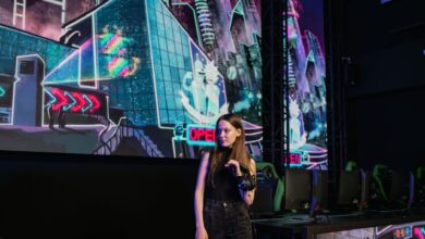 A woman stands in a virtual reality gaming arena with vibrant neon backdrops, wearing casual attire.