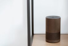 A sleek, modern smart speaker displayed on a wooden shelf in a minimalist room setting.