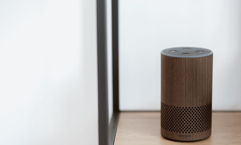 A sleek, modern smart speaker displayed on a wooden shelf in a minimalist room setting.