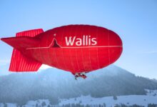 Bright red airship flying over snowy mountains in Château-d’Œx, Switzerland, with clear blue skies.