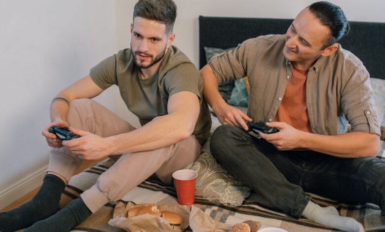 Two friends sitting on a bed, playing video games and enjoying snacks indoors.