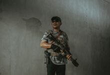 A soldier wearing tactical gear and sunglasses holds an assault rifle indoors.