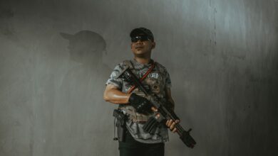 A soldier wearing tactical gear and sunglasses holds an assault rifle indoors.