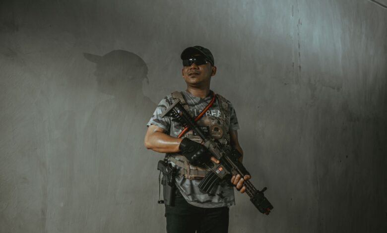 A soldier wearing tactical gear and sunglasses holds an assault rifle indoors.