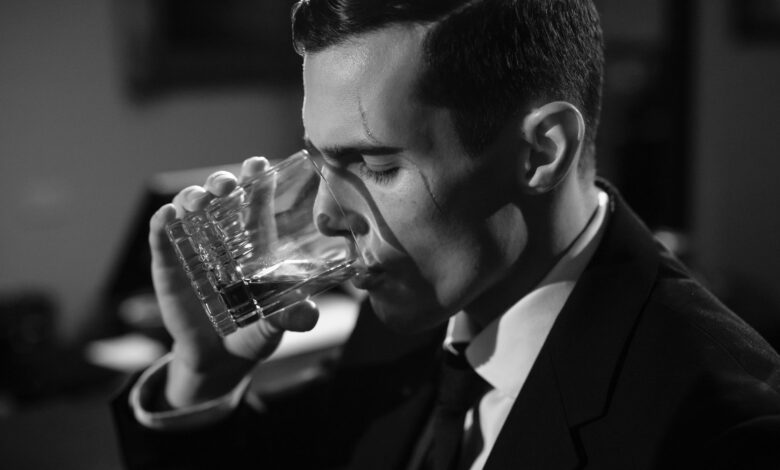 Black and white image of a man sipping whiskey, evoking classic film noir style.