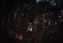 Anonymous creepy ghost woman in white bloody dress walking in dark cemetery near old rusty metal gates