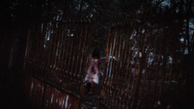 Anonymous creepy ghost woman in white bloody dress walking in dark cemetery near old rusty metal gates