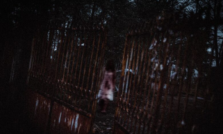 Anonymous creepy ghost woman in white bloody dress walking in dark cemetery near old rusty metal gates