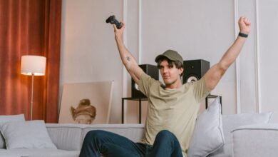 A young man joyfully celebrates winning a video game, sitting on a couch with his arms raised in triumph.