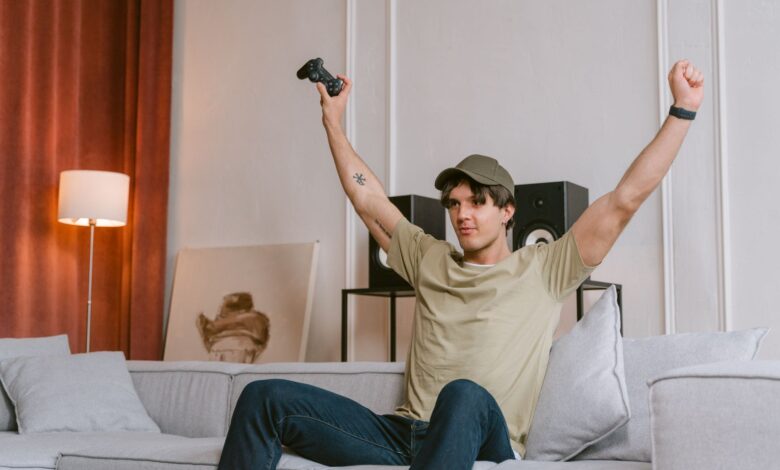 A young man joyfully celebrates winning a video game, sitting on a couch with his arms raised in triumph.