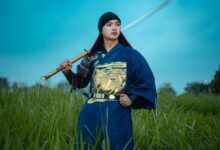 A samurai in traditional attire holds a sword in a grassy field during twilight.