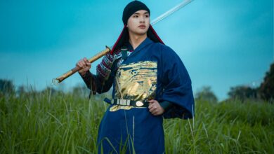 A samurai in traditional attire holds a sword in a grassy field during twilight.