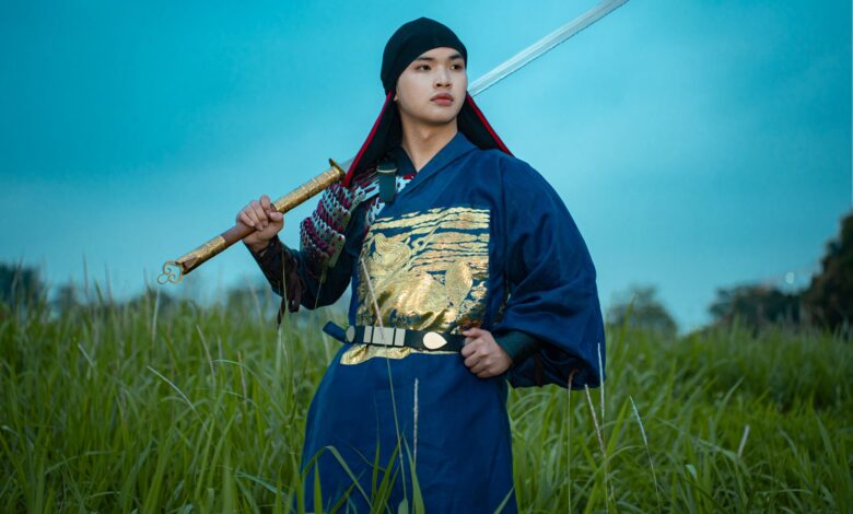 A samurai in traditional attire holds a sword in a grassy field during twilight.