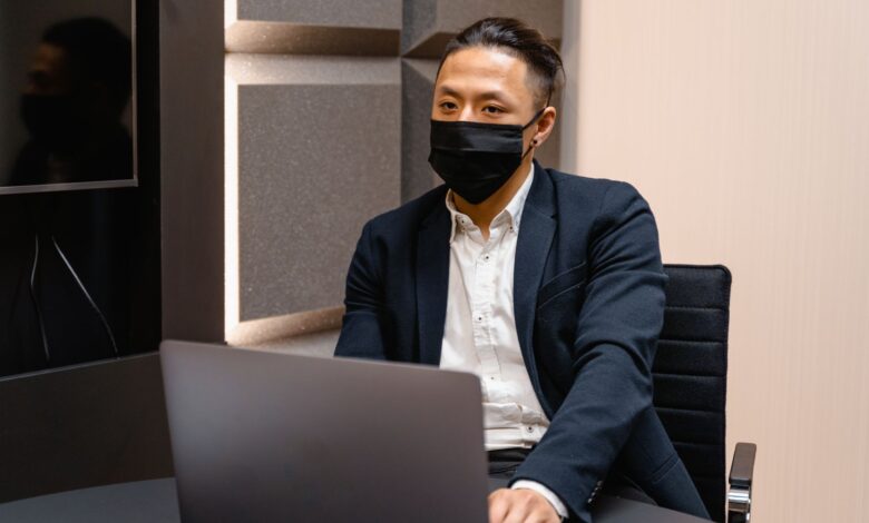 Professional businessman in formal attire wearing a face mask while working on a laptop in an office.