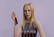Blonde woman holding a vial with red liquid, set against a purple background.