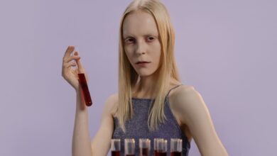 Blonde woman holding a vial with red liquid, set against a purple background.