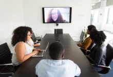 From above of diverse employees gathering at table in contemporary conference hall and having video call with coworker on TV set