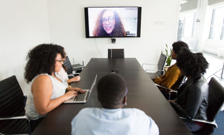 From above of diverse employees gathering at table in contemporary conference hall and having video call with coworker on TV set