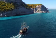 Drone view of aged sail ship floating in rippling blue ocean near rocky mountainous island with green trees and plants under blue cloudless sky in sunny summer day