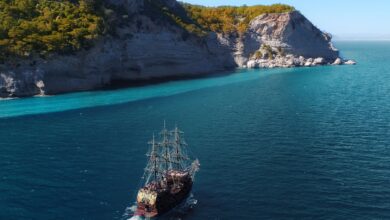 Drone view of aged sail ship floating in rippling blue ocean near rocky mountainous island with green trees and plants under blue cloudless sky in sunny summer day