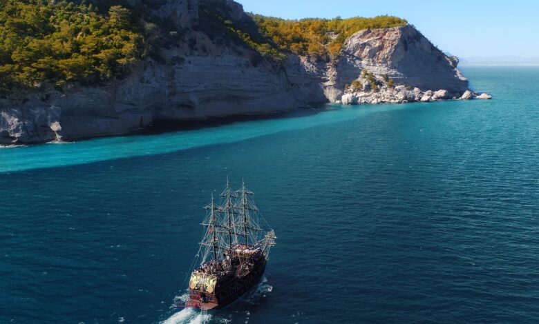 Drone view of aged sail ship floating in rippling blue ocean near rocky mountainous island with green trees and plants under blue cloudless sky in sunny summer day