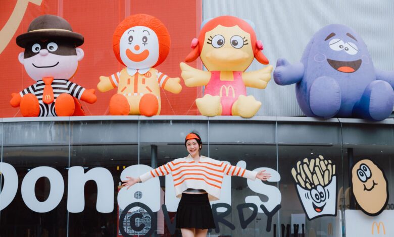 Woman posing happily in front of colorful inflatable fast food characters, radiating playful energy.