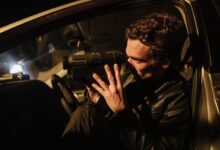 A man in a leather jacket uses a camcorder at night inside a car.