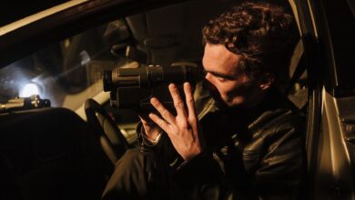 A man in a leather jacket uses a camcorder at night inside a car.
