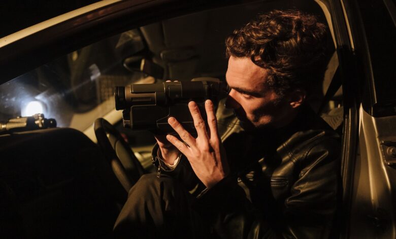 A man in a leather jacket uses a camcorder at night inside a car.