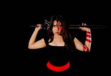Fierce female warrior in cosplay with sword and face paint, poised against a black background.