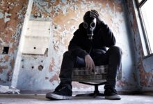 A man wearing a gas mask is seated in a dilapidated room, conveying a sense of despair and survival.
