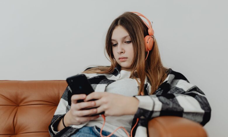 Teenage girl sitting on a sofa listening to music on headphones and using a smartphone.