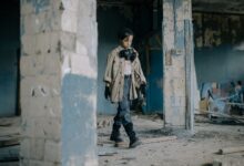 A young child walks in a post-apocalyptic, abandoned building wearing a gas mask.