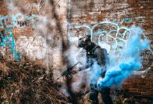 A person in tactical gear engages in an airsoft game with colorful smoke in an urban graffiti area.