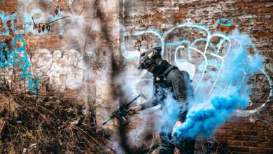 A person in tactical gear engages in an airsoft game with colorful smoke in an urban graffiti area.