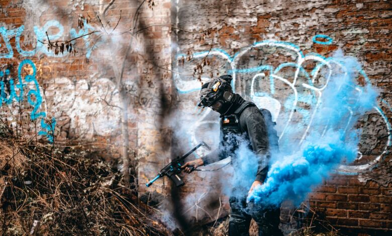 A person in tactical gear engages in an airsoft game with colorful smoke in an urban graffiti area.