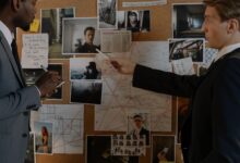 Two detectives analyze a crime board filled with photos, maps, and notes for an investigation.