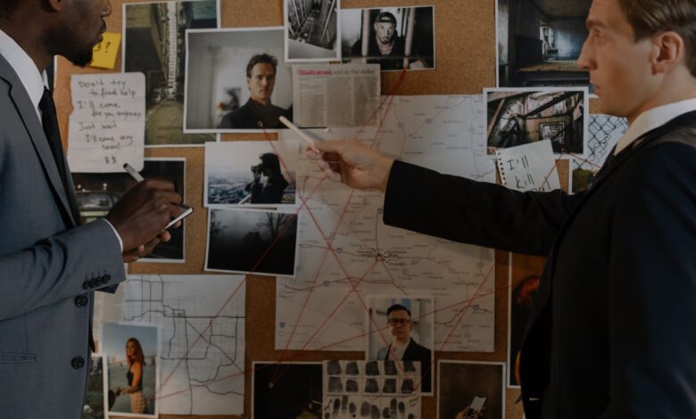 Two detectives analyze a crime board filled with photos, maps, and notes for an investigation.