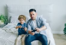 Father and son joyfully playing video games while sitting on a bed, capturing a moment of family fun.